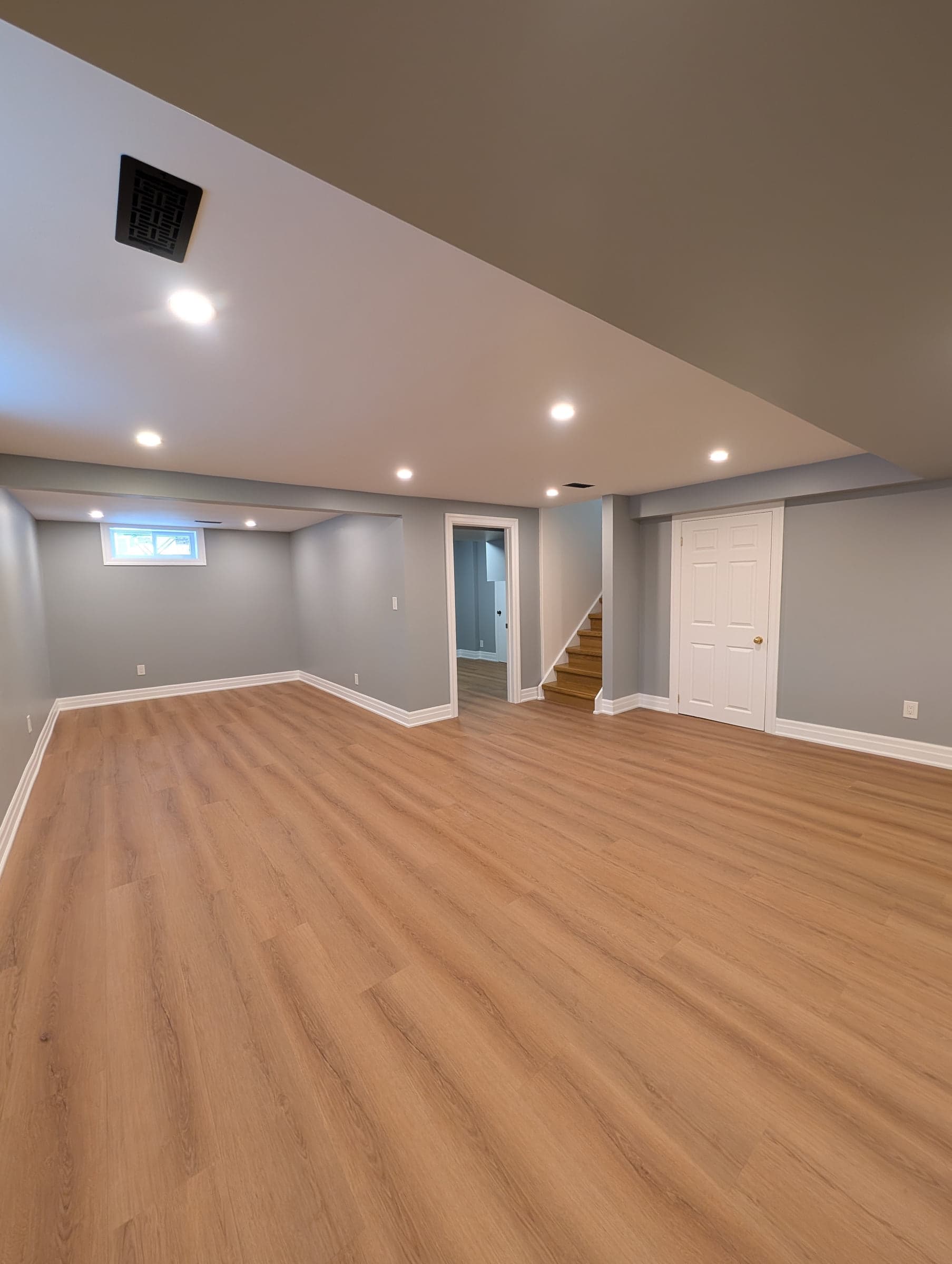 Finished basement room with grey walls, light oak vinyl plank flooring, recessed lighting, a white door, and stairs up to the main floor