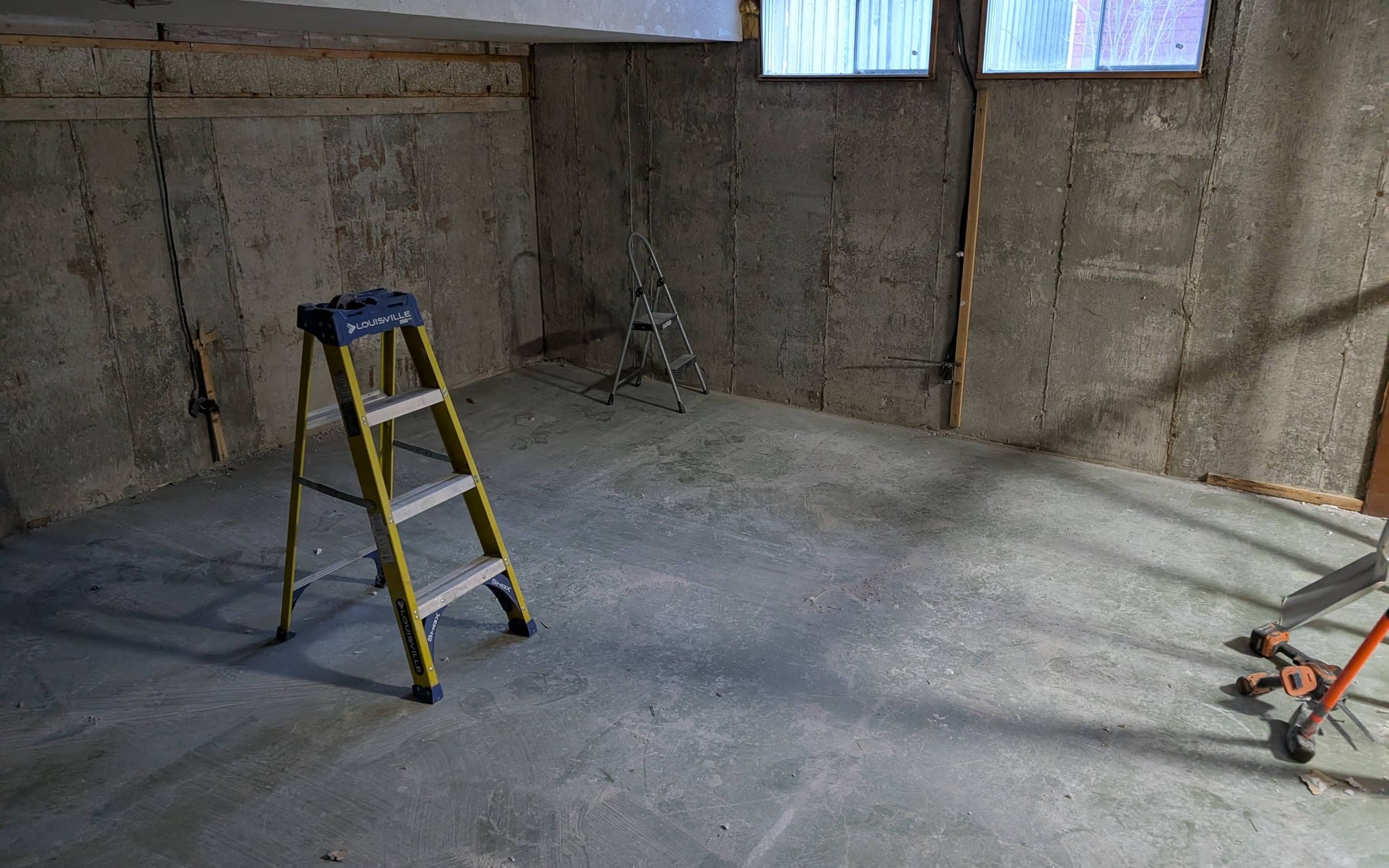 Unfinished basement with bare concrete walls, exposed floor joists, two small hopper windows, and a stepladder on the concrete floor