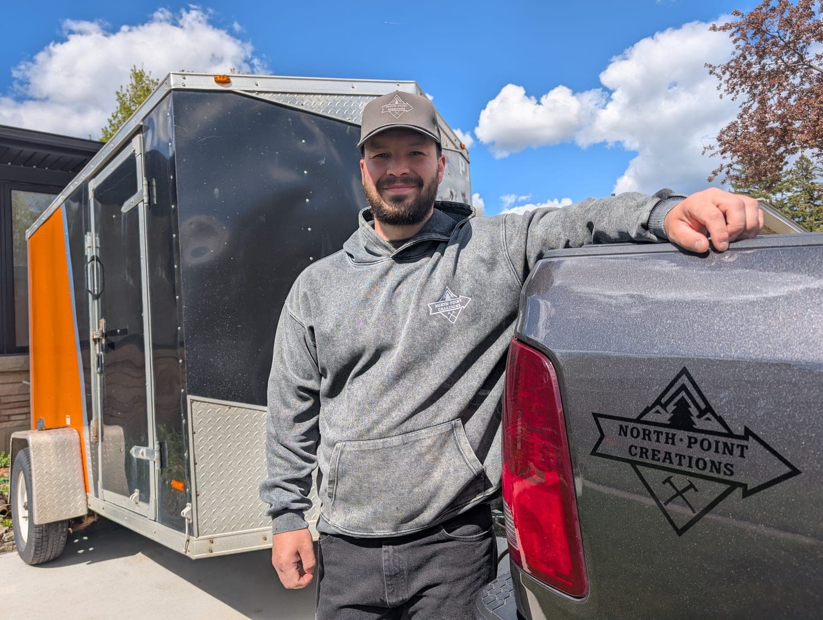 Kennedy in a North Point Creations hoodie and cap leaning on the tailgate of a logo-branded pickup truck, with the company's enclosed cargo trailer parked behind him on a sunny driveway