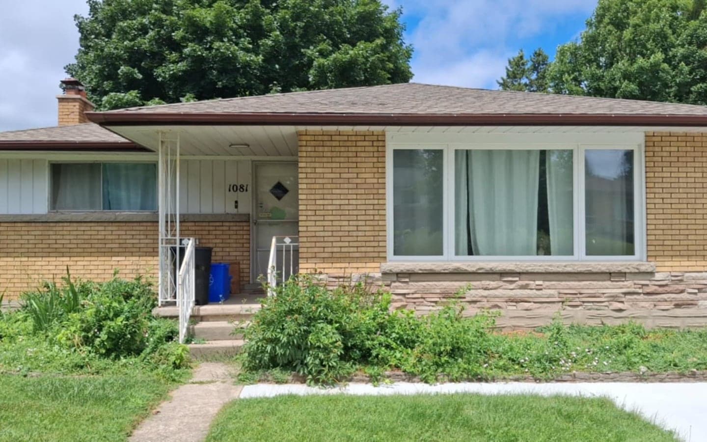 Yellow brick bungalow before renovation, with an overgrown front garden bed, thin white metal porch railings, and a cracked concrete walkway to the front door