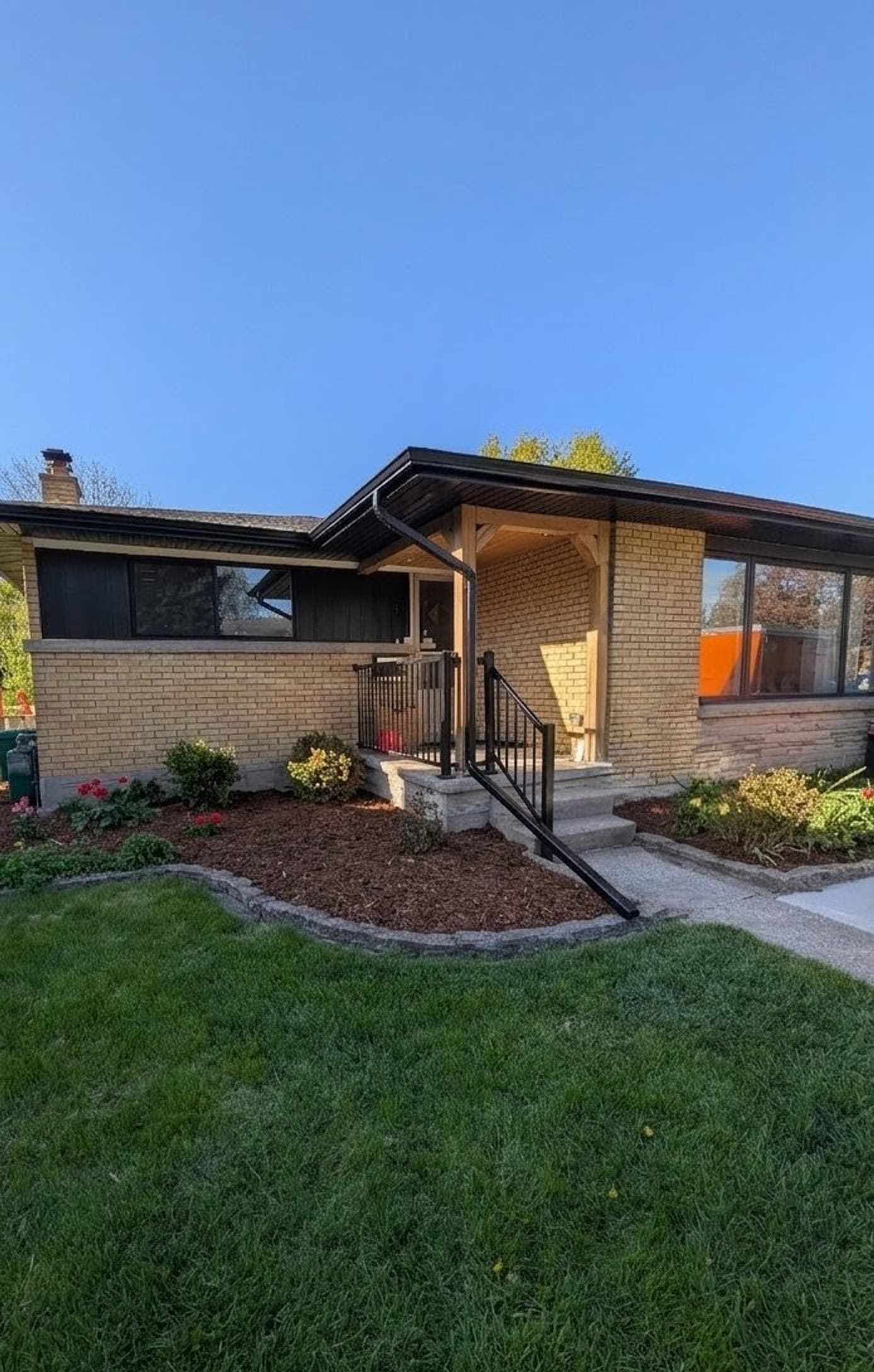 Renovated yellow-brick bungalow with a timber post-and-beam entry canopy, landscaped garden beds, and fresh sod