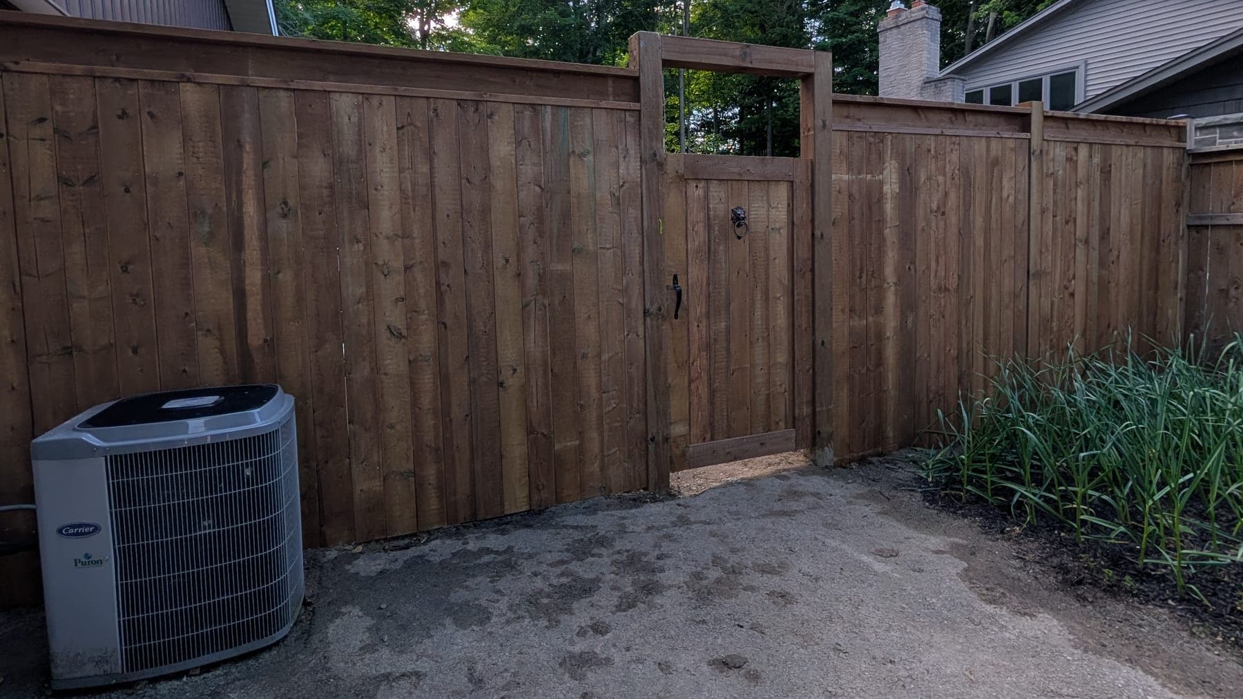 Replacement brown-stained board fence and matching gate hung in an open square arbour frame with black ring-pull hardware, above a levelled gravel side yard