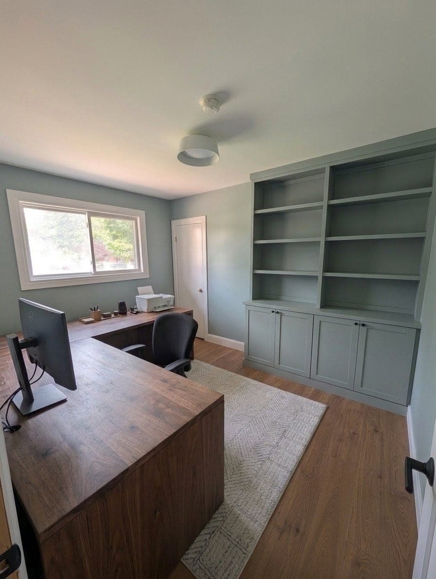 Home office with an L-shaped walnut desk, monitor and printer, and floor-to-ceiling sage-green built-in shelving and cabinets