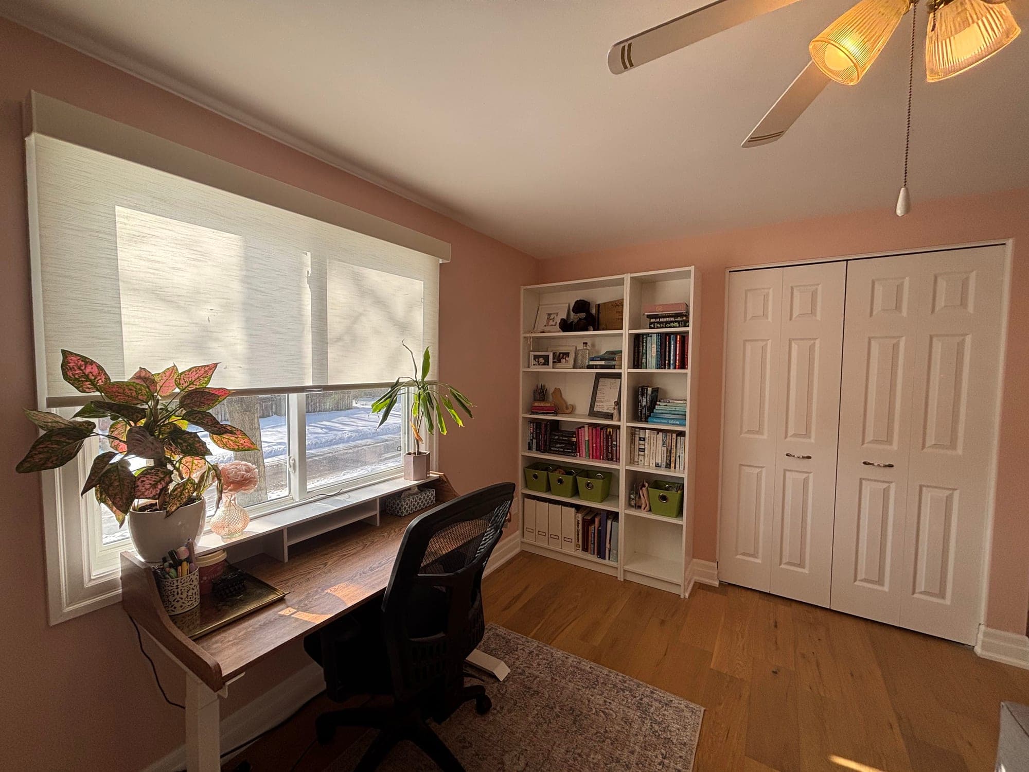 Home office with a wood desk and mesh task chair beneath a wide shaded window, white bookshelves holding books and storage bins, and a white closet door