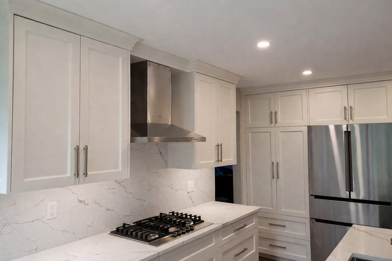 White shaker cabinetry around a gas cooktop, range hood, and stainless refrigerator